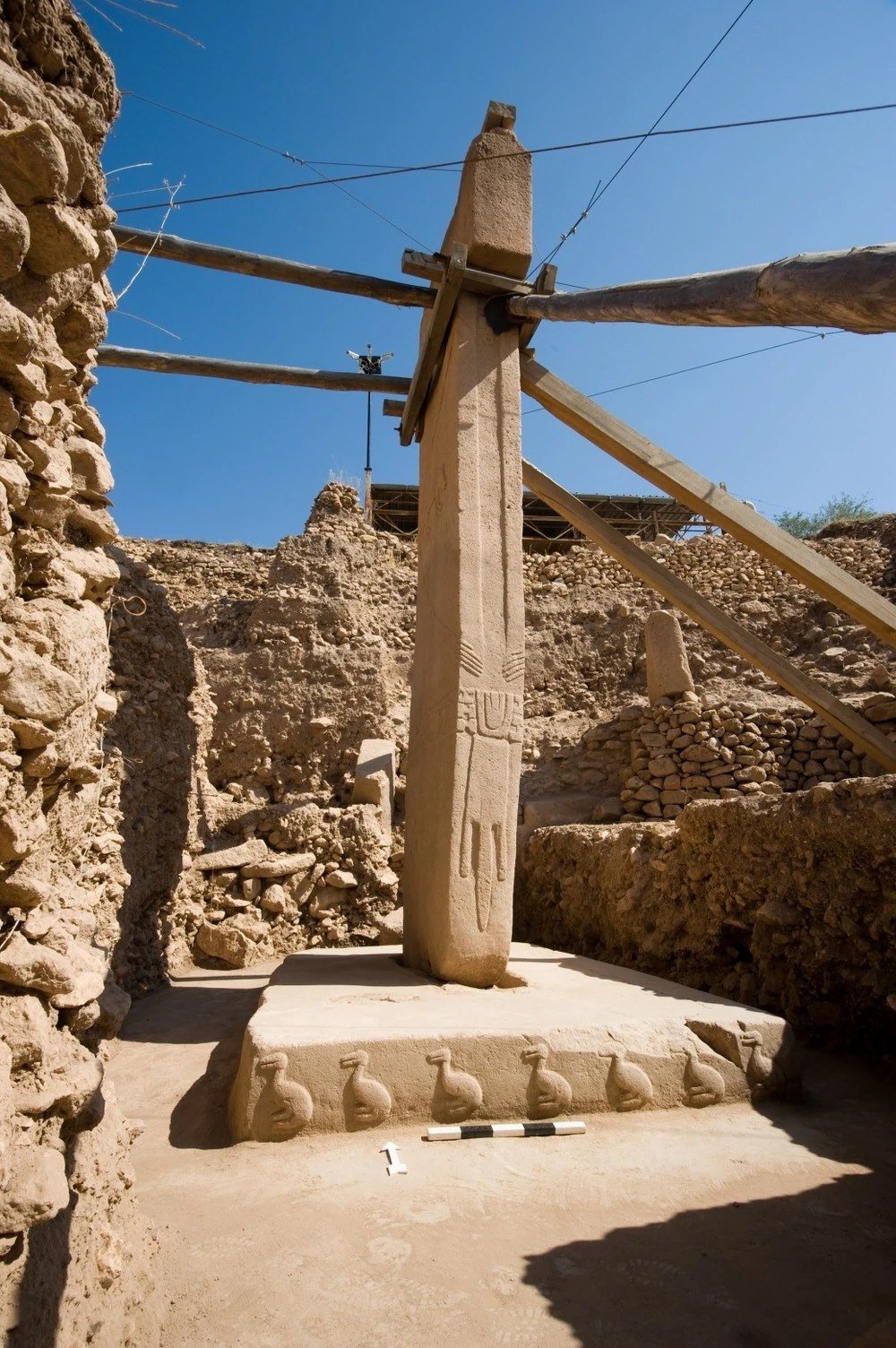 A tall central T-pillar of Enclosure D on a plastered pedestal, with low-relief human arms, hands and a belt on the shaft and a row of carved ducks along the pedestal base