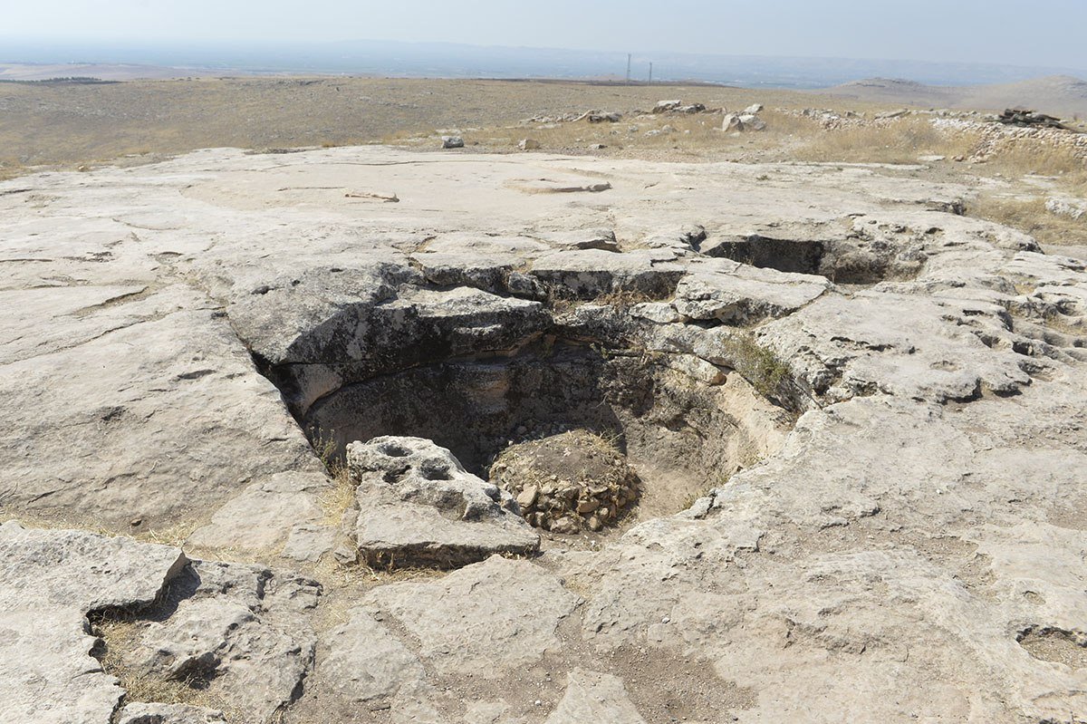 A large basin cut into flat limestone bedrock on the hilltop, with the wide landscape behind
